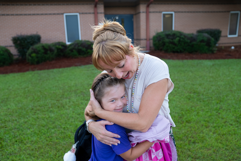 Woman embracing a young girl with a backpack on a lawn in front of a building.