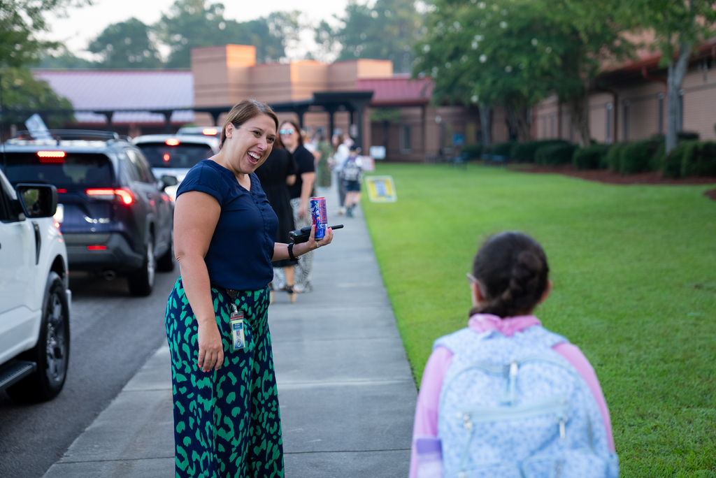 Woman in blue top and green skirt smiles while holding can, standing on sidewalk; girl with pink top and backpack behind her.