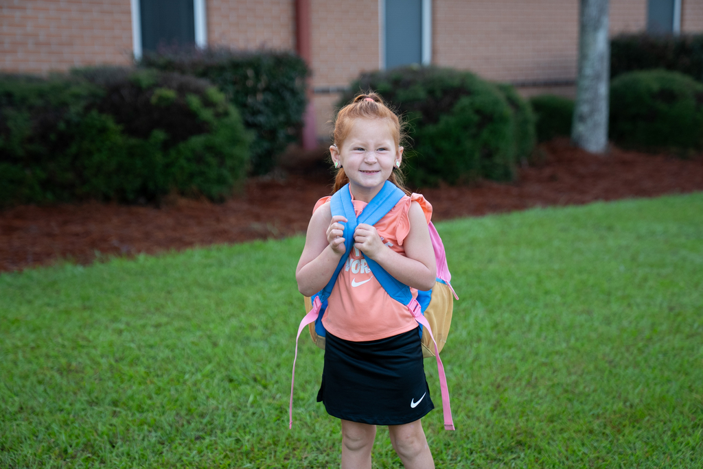 A young child with red hair, standing in grass, wearing a backpack. Behind the child is a brick building.