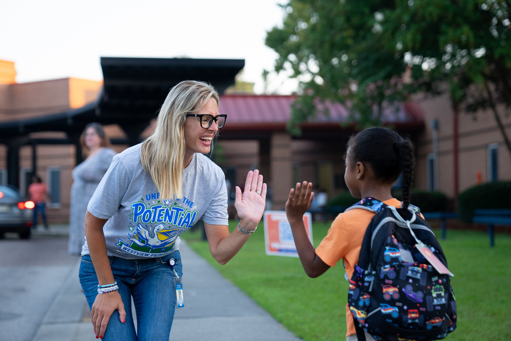 Woman in glasses and a gray shirt greets a child with a high-five. Child wears backpack and orange shirt.