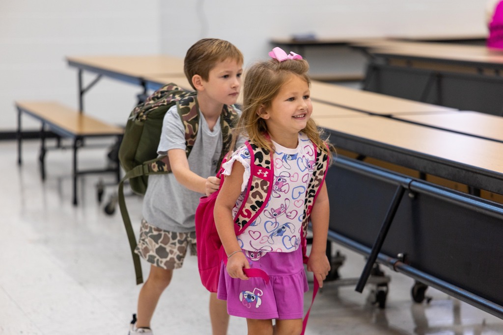 Two children walking in a classroom; a boy with a backpack and a girl with a pink bag.
