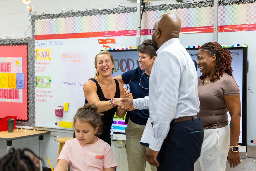 Group of adults and a child in a classroom. A man in white shakes hands with a smiling woman in black.
