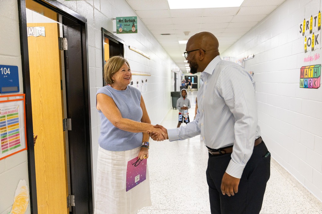 Two adults, one in a white shirt and the other in a blue shirt, shake hands in a school hallway.