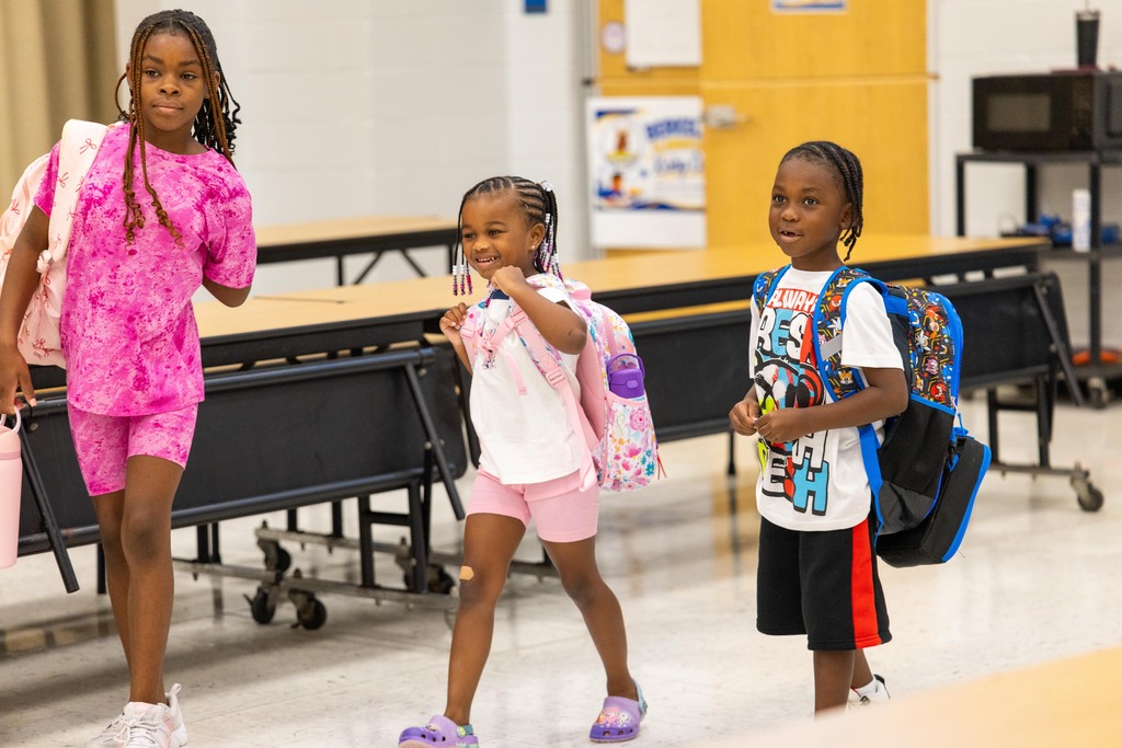 Three children in a classroom, each carrying a backpack, walking through a room with tables and chairs.