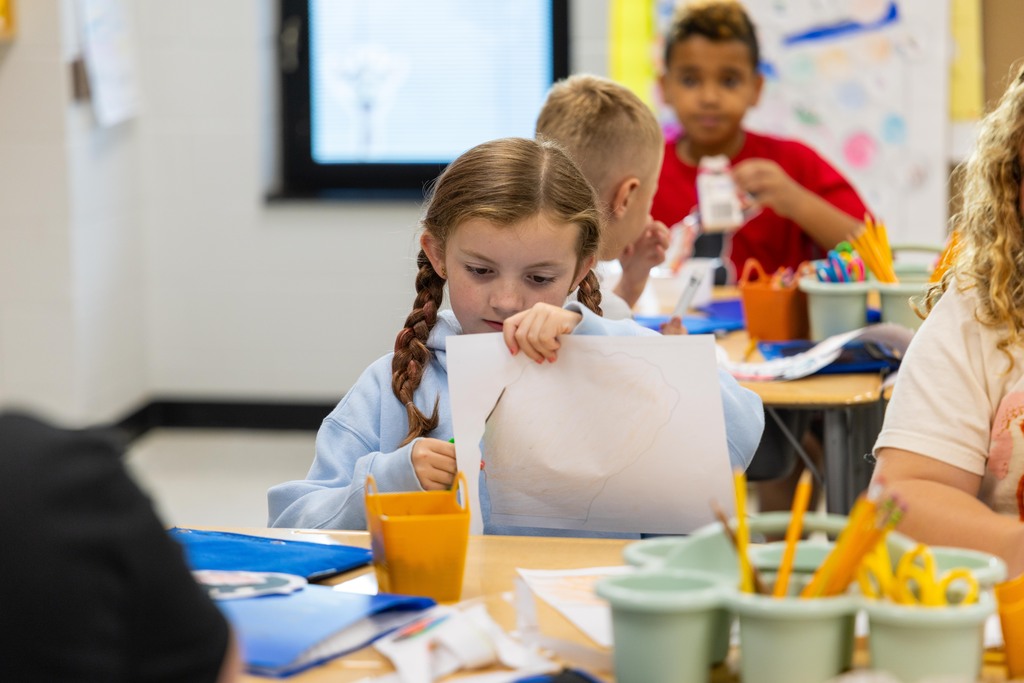 Students in a classroom. One girl focuses on paper, holding it with both hands. Nearby, various school supplies are on the table.