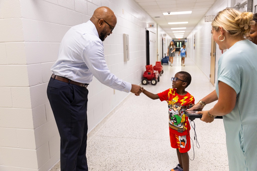 A man shakes hands with a young boy. Behind them, a woman holds a small child. The hallway has white walls and a yellow line on the floor.