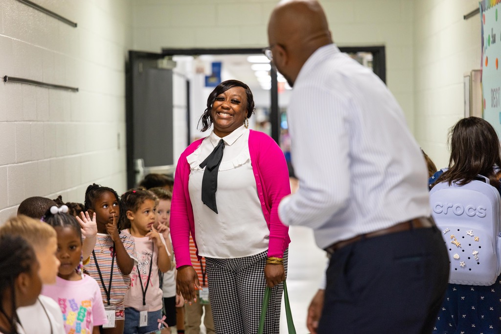 Two adults, one in pink, one in white, and a belt, stand with children in a hallway.