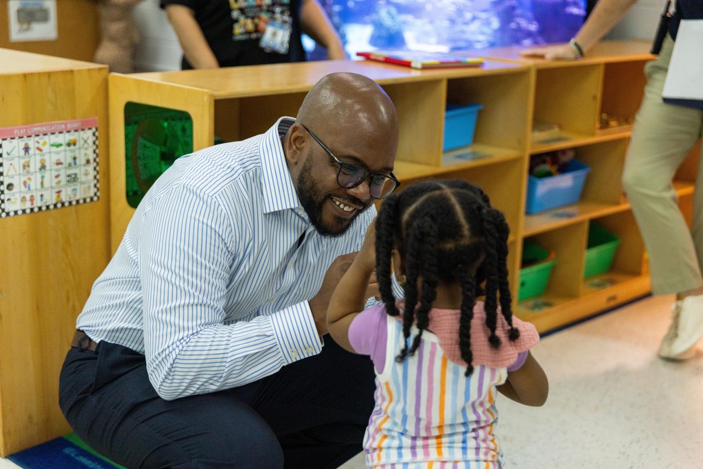 A man with glasses crouches in front of a young girl, who smiles, in a classroom.