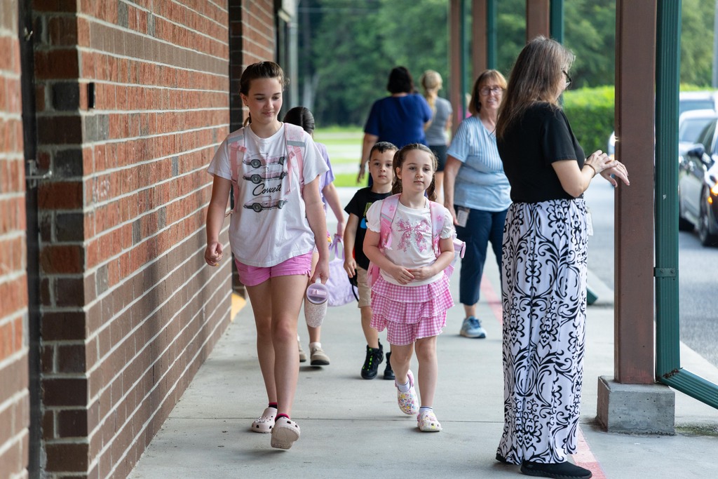 Several children and adults walk along a sidewalk near a brick building with trees in the background.