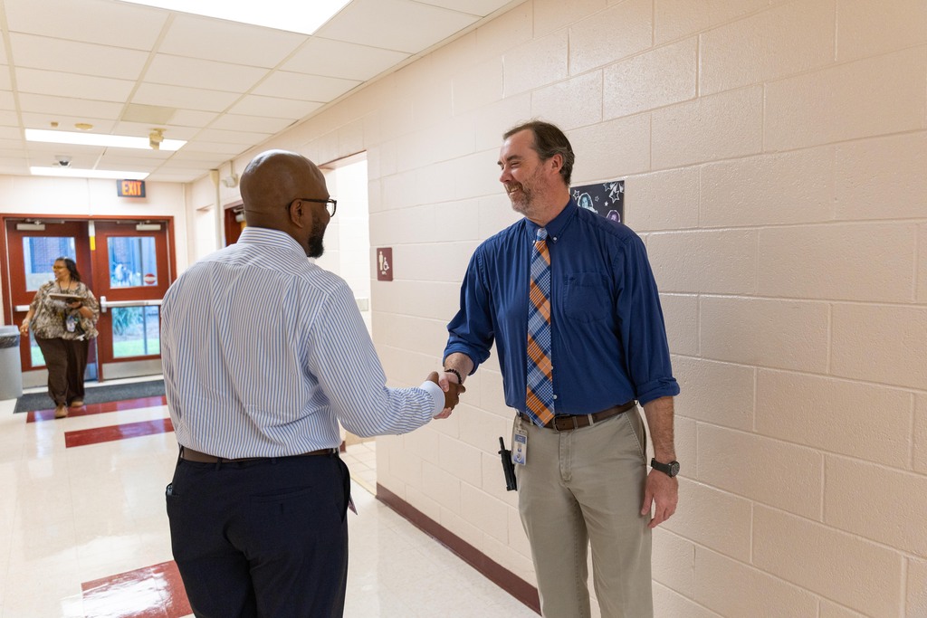 Two men shake hands in a hallway. One wears a blue shirt and tie; the other wears a white shirt. Background shows a hallway with doors and a trash can.