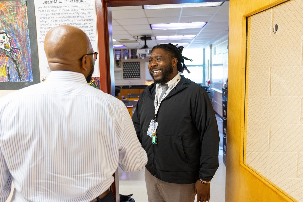 Two men stand in a hallway, one in a white shirt, the other in a black jacket. They appear to be in conversation. An open door is behind them.