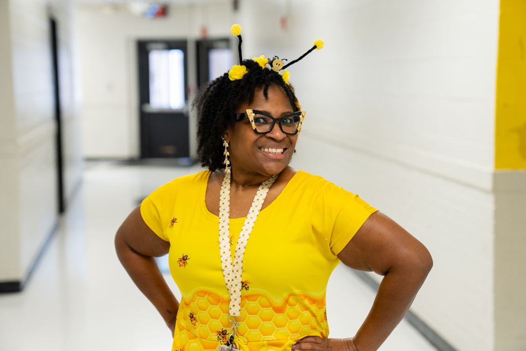 A woman in a yellow bee costume stands in a hallway with a yellow wall, smiling.