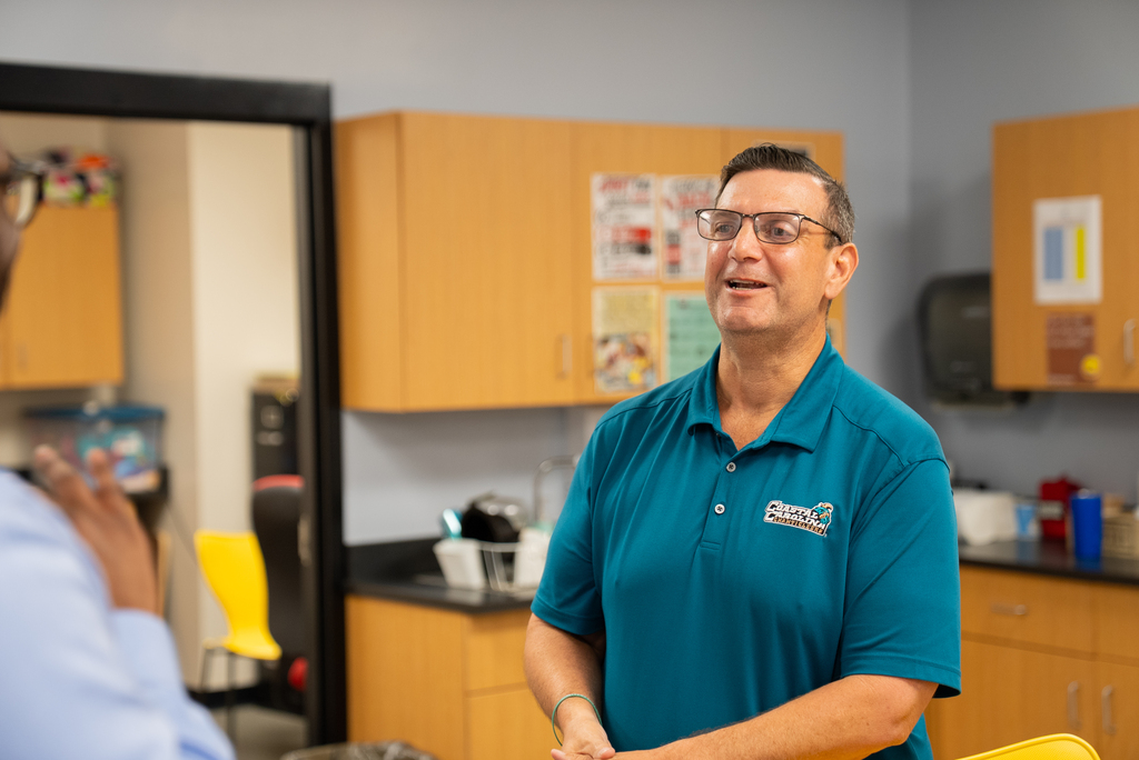 A man in a teal shirt with glasses speaks to another person in a classroom. Behind them are cabinets and a trash bin.