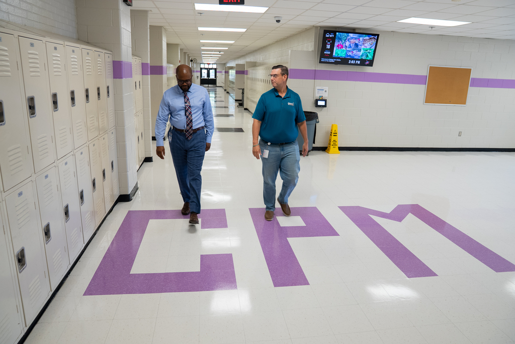Two men walk down a school hallway, past lockers, and over large letters on the floor that spell "CFM."