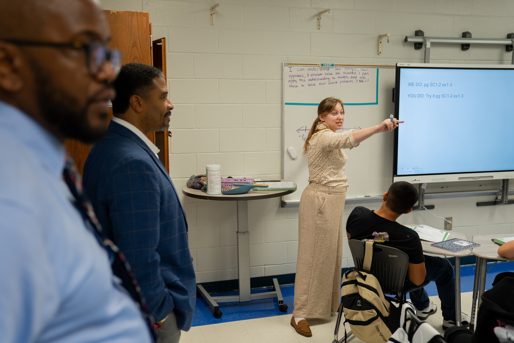 A teacher points to a screen while two men and a student listen. Behind them, a white board and a round table are visible.