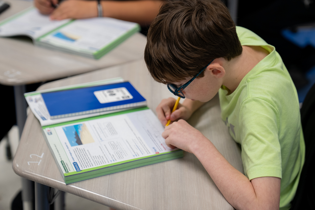 A boy sits at a desk, wearing glasses and a green shirt, writing in a notebook.