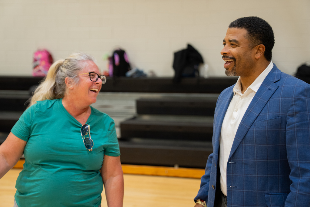 A man in a blue jacket and a woman in a green shirt are smiling and talking in a gym.