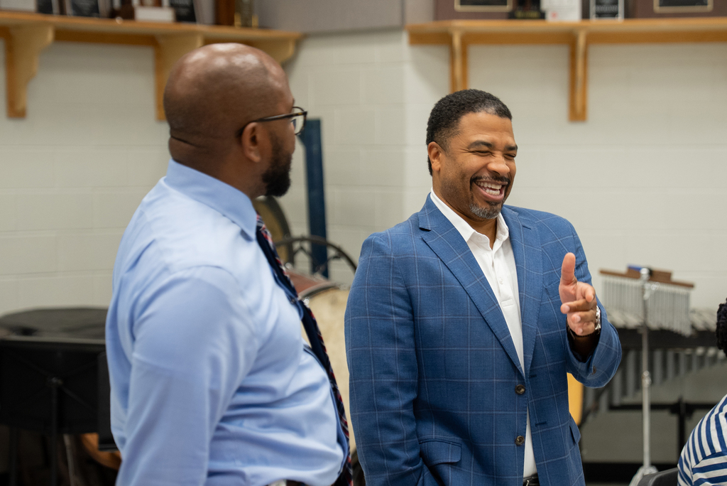 Two men in suits and ties smile and speak in a music classroom with shelves and musical instruments.