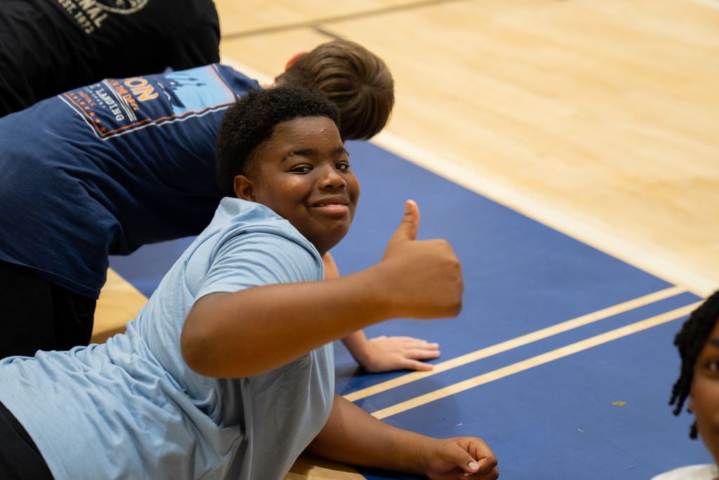 A boy smiles and gives a thumbs-up gesture while lying on a blue mat on a wooden floor.