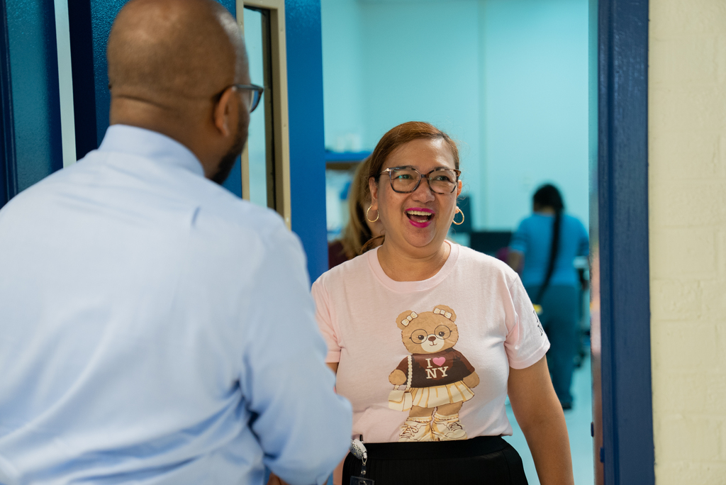 A person with glasses talks to a smiling woman in a pink shirt with a bear design. Behind them, another person in a white shirt and black skirt walks.