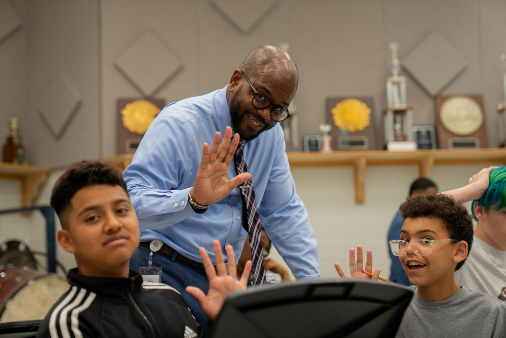 A man stands in a classroom, gesturing with his hands while three students look up at him.