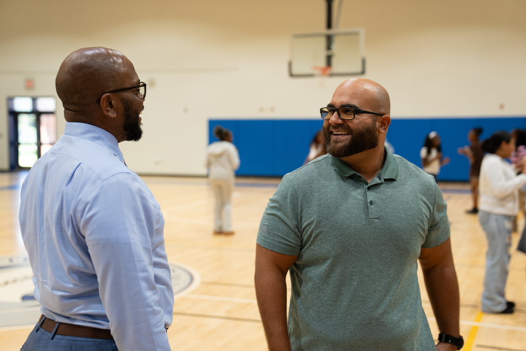 Two men in a gym, one wearing a blue shirt and glasses, the other in a green polo, talking.