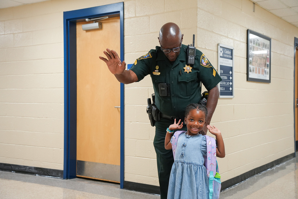A police officer stands next to a young girl with a backpack, both making hand gestures in a hallway.