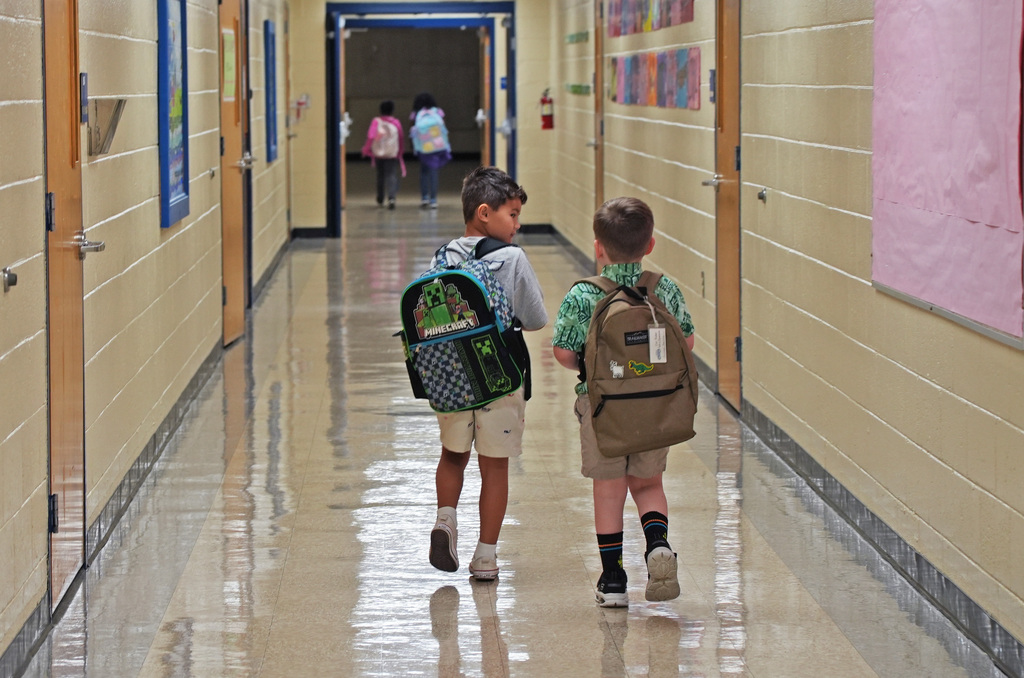 Two children in a hallway with bright light. One wears a green backpack and shorts; the other wears a blue backpack and socks.