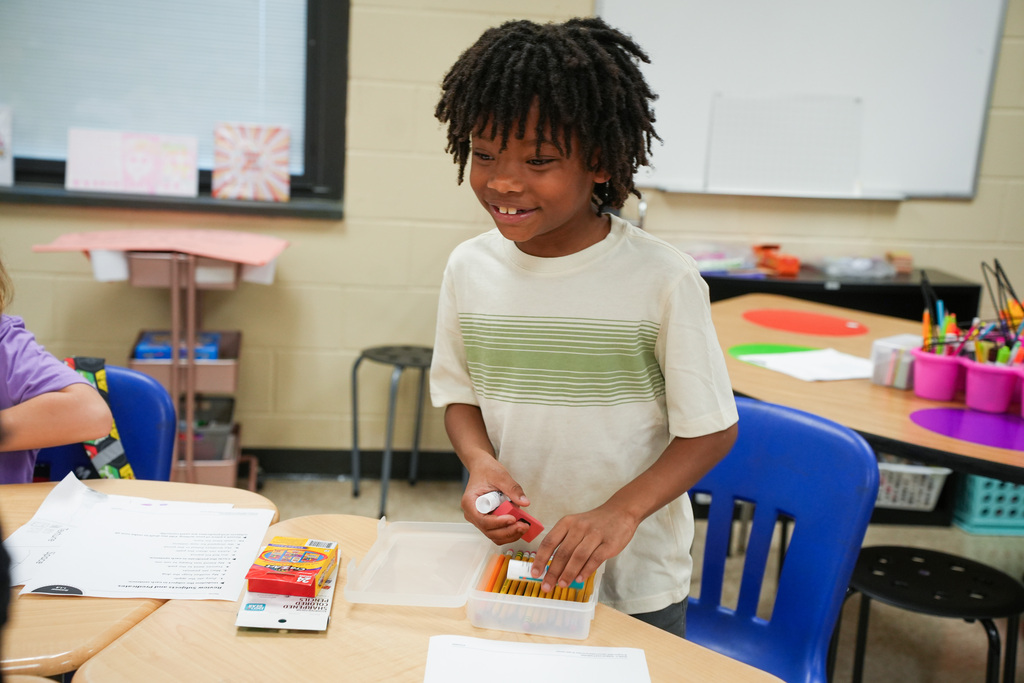 A child is standing at a desk in a classroom, holding a box of crayons. Behind them, a whiteboard and tables with school supplies are visible.
