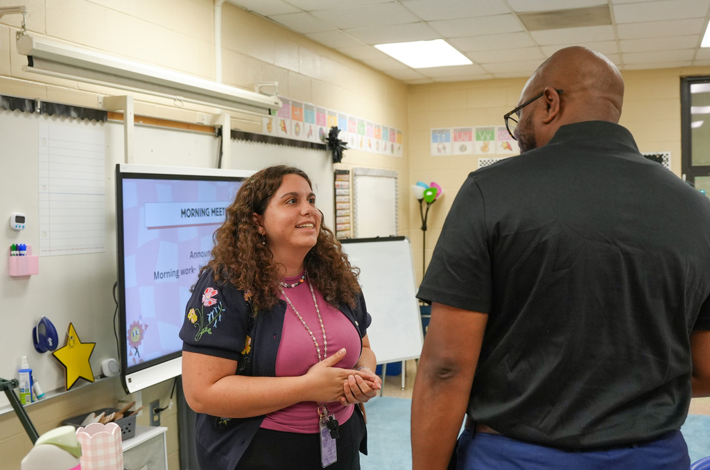 Two adults facing each other in a classroom. One wears a pink top and has curly hair, the other wears a black shirt and glasses.