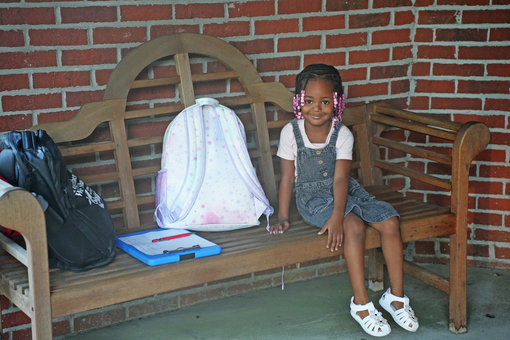 A young girl sits on a wooden bench with a brick wall behind her. She has a backpack and a folder on the bench.