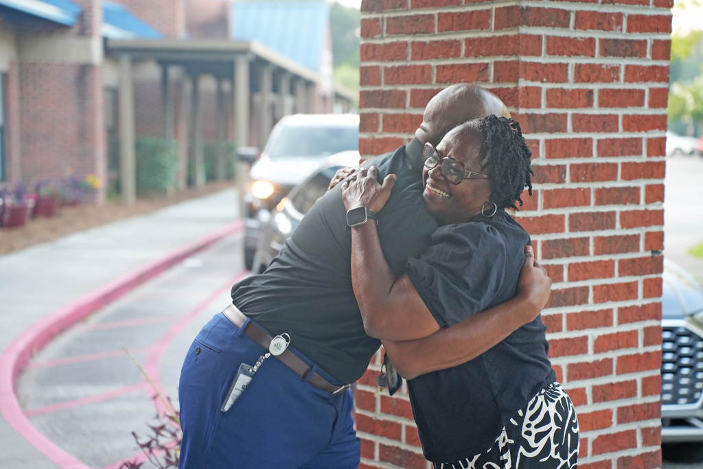 Two individuals embrace tightly next to a brick column. The man wears a black shirt, belt, and watch. The woman wears a patterned skirt. A car is parked behind them.