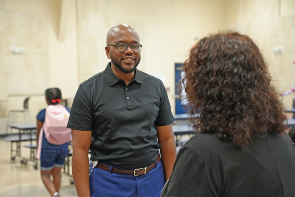 Two individuals in a school cafeteria. A man in a black shirt and blue pants stands. A woman in a black shirt has curly hair.