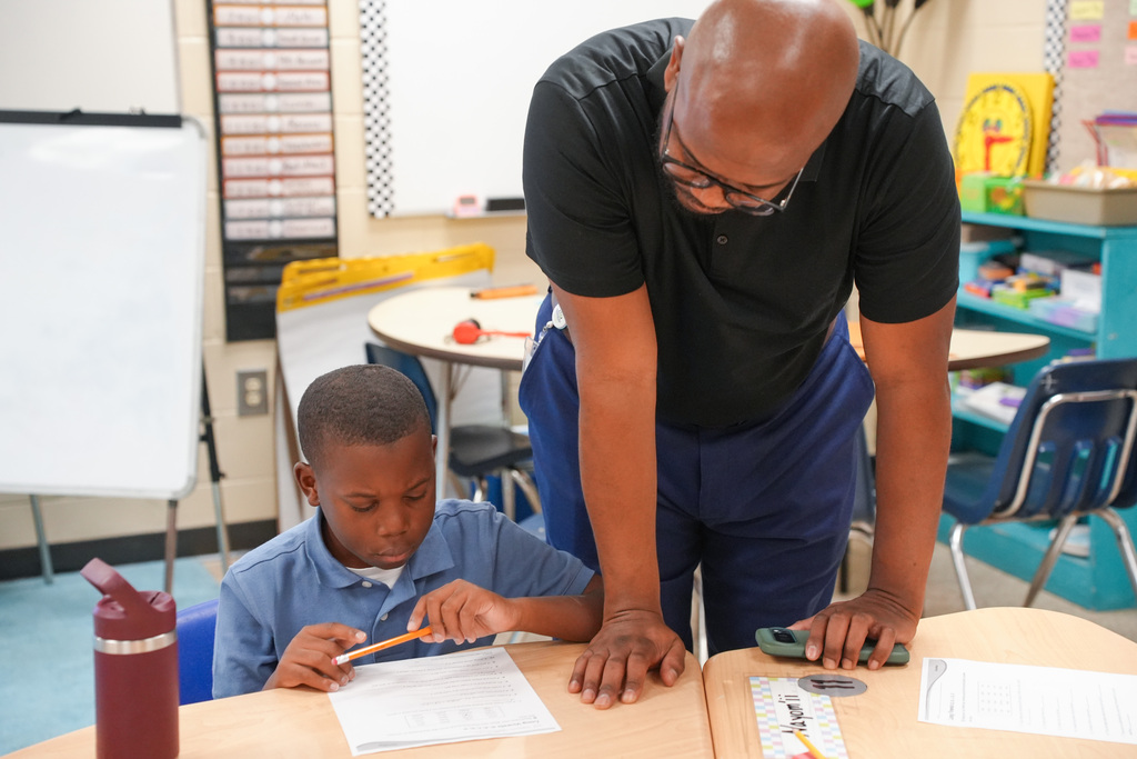 A man helps a boy seated at a desk, they are both holding pencils and looking at a paper.