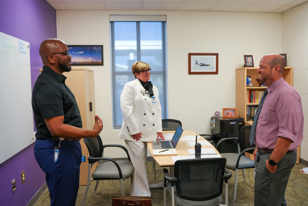 Three people are in a meeting room with a table and chairs; one person stands in front of a laptop.