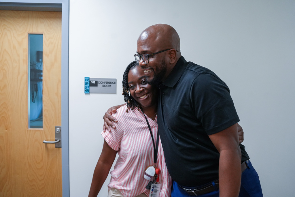 A man and woman stand close, smiling and hugging in an indoor setting. The man wears glasses and a black shirt.