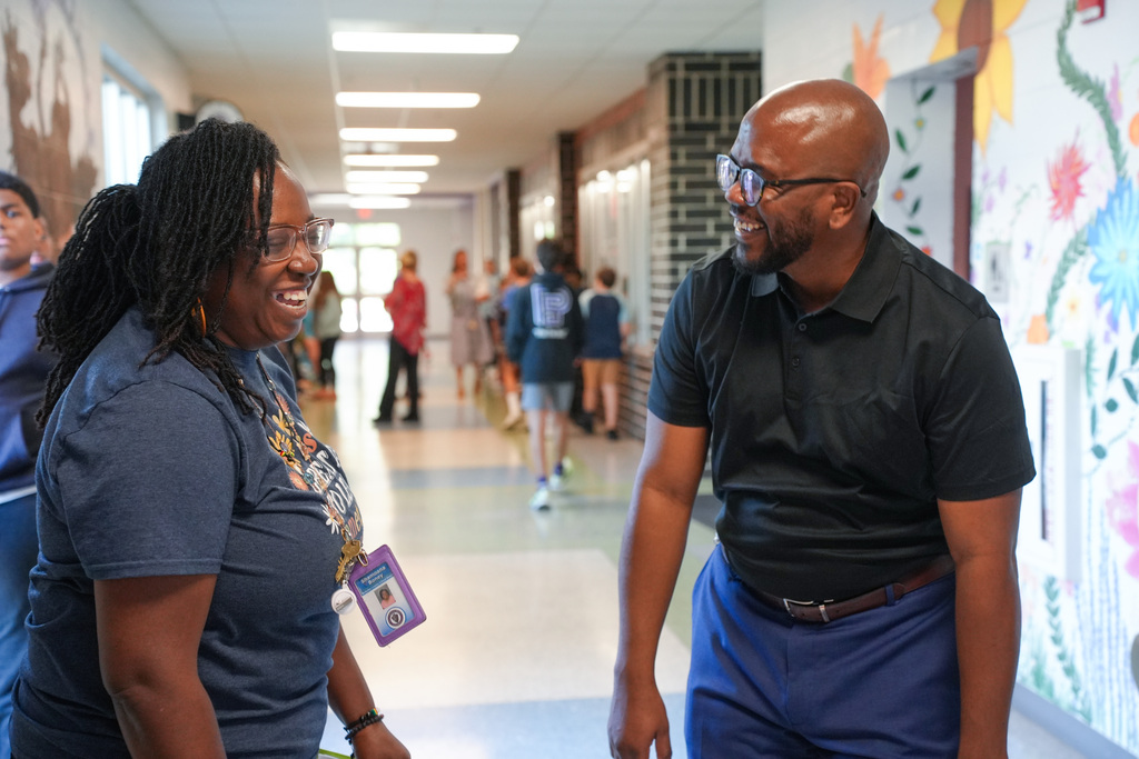 Two adults engage in conversation within a hallway; one wears a blue shirt, the other a black shirt. Background shows a colorful wall, people, and a bright corridor.