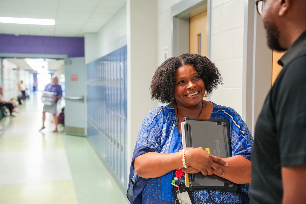 A woman with curly hair smiles and holds a folder while standing in a hallway.