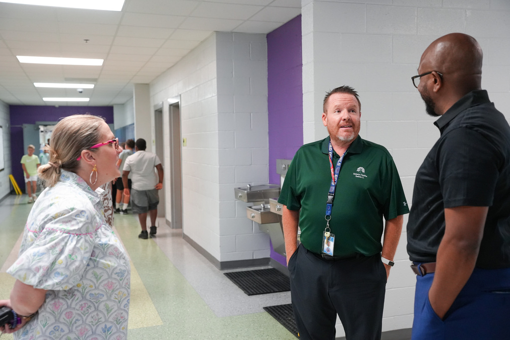 Three adults converse in a school hallway with white and purple walls. A woman in glasses and a floral shirt stands on the left.
