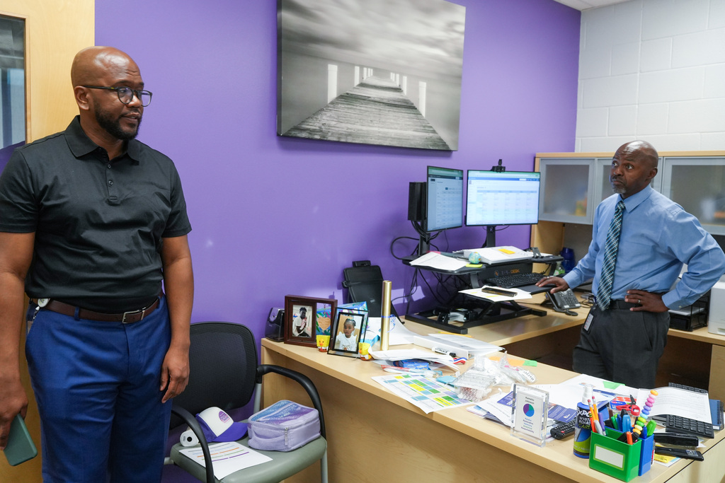 Two men in a purple-walled office. One man stands at a desk with monitors and papers; the other stands in front of him.