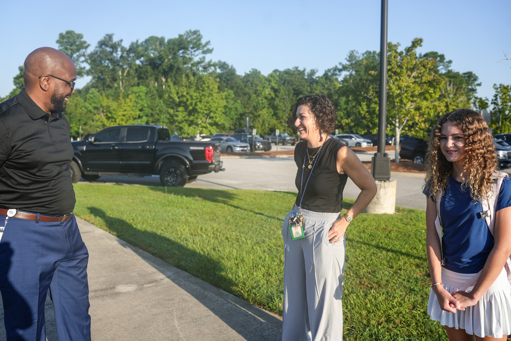 A man and two women converse in a parking lot. The man wears a black shirt and blue pants. Both women wear black tops.