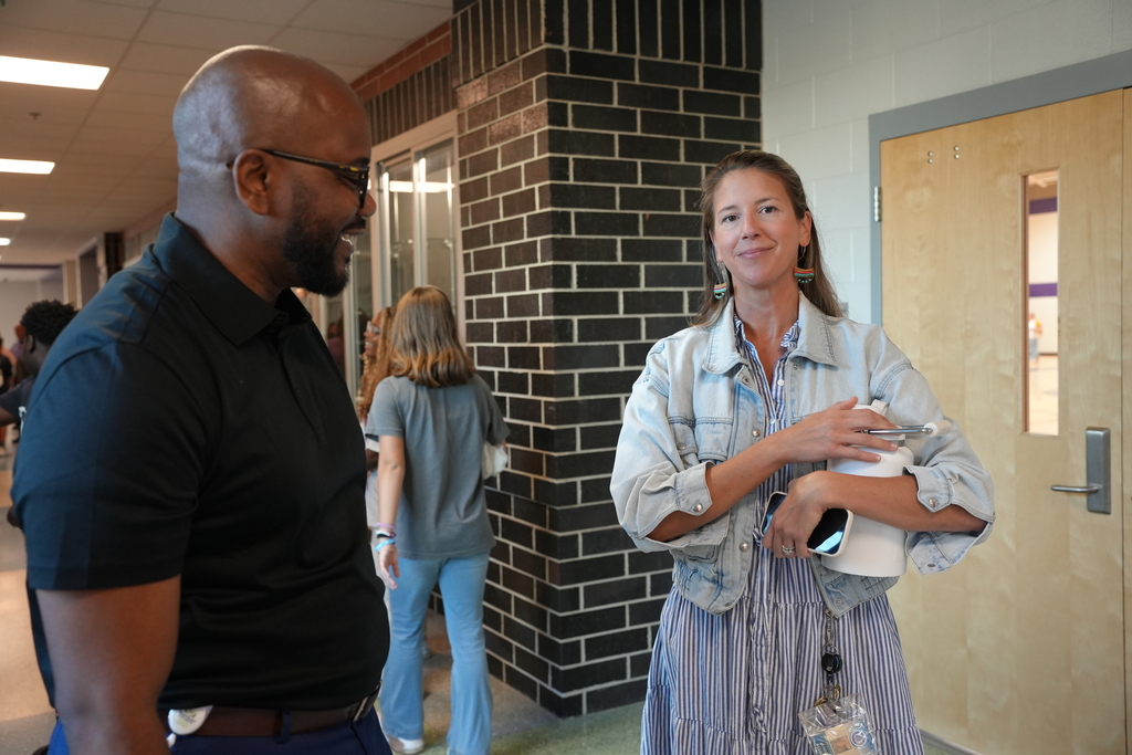 Two people conversing inside a corridor. One wears a black shirt and glasses; the other wears a denim jacket and holds a bottle. Behind them, a person walks in the background.