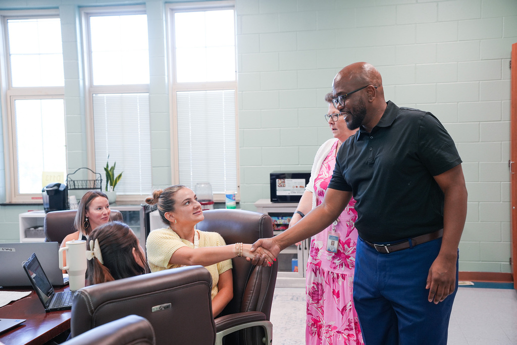A group of people in a room, a man shakes hands with a woman in a yellow shirt.