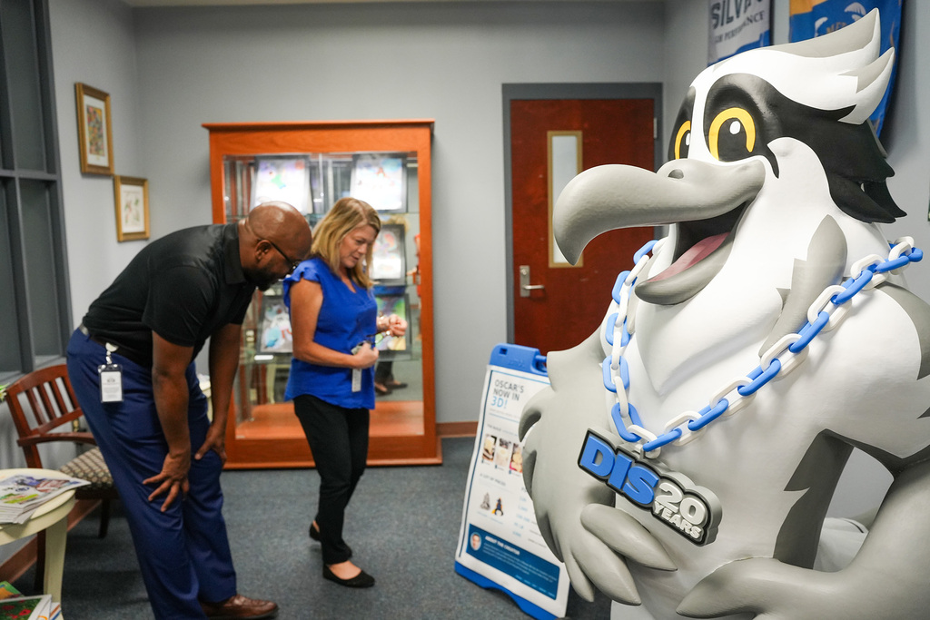 A man and a woman are looking at a mascot with the word "DISC" on it. The room has wooden furniture.