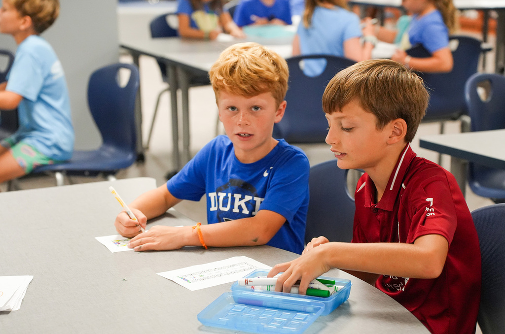 Two boys in a classroom, one writing and another using a calculator. Behind them, more children are seated at tables.