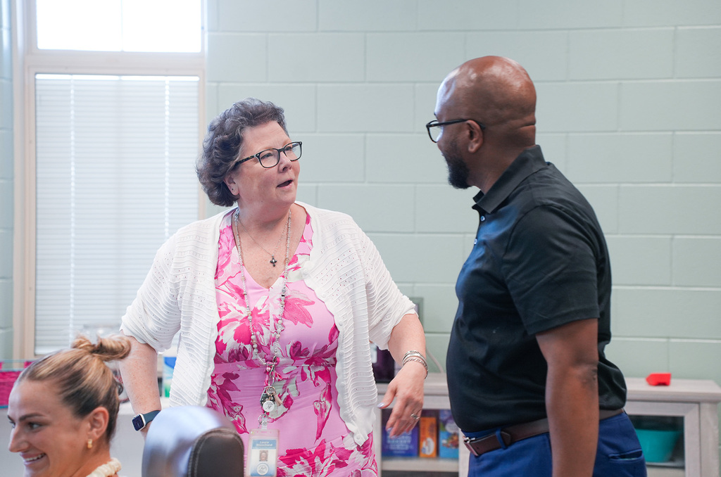 A woman in glasses stands in front of a man in a black shirt and blue pants. Behind them is a woman with a ponytail.