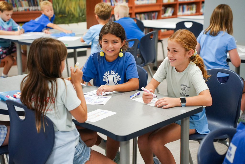 Three girls sit at a table in a classroom. One holds a marker and paper, another smiles, and the third listens attentively.
