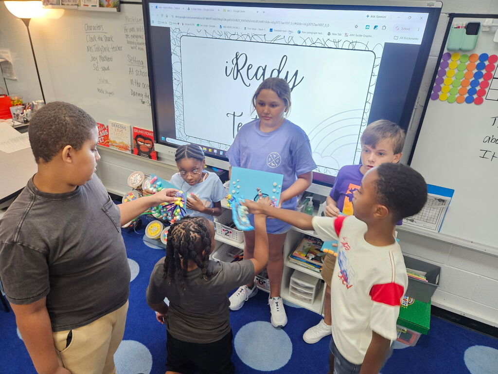 Kids stand in a circle around a teacher in a classroom. They hold a book as the teacher shows something on a monitor.