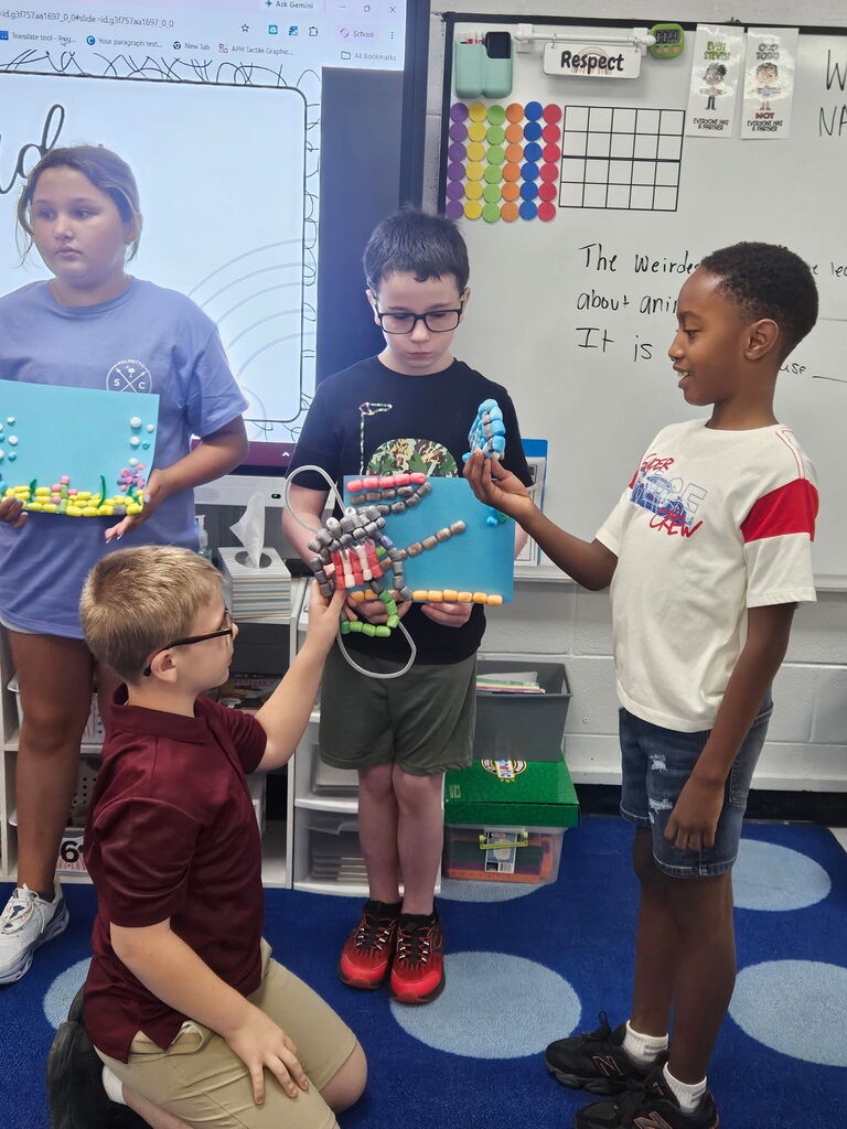 Four children are standing in a classroom. One child kneels, holding a mechanical object, and others look at it.