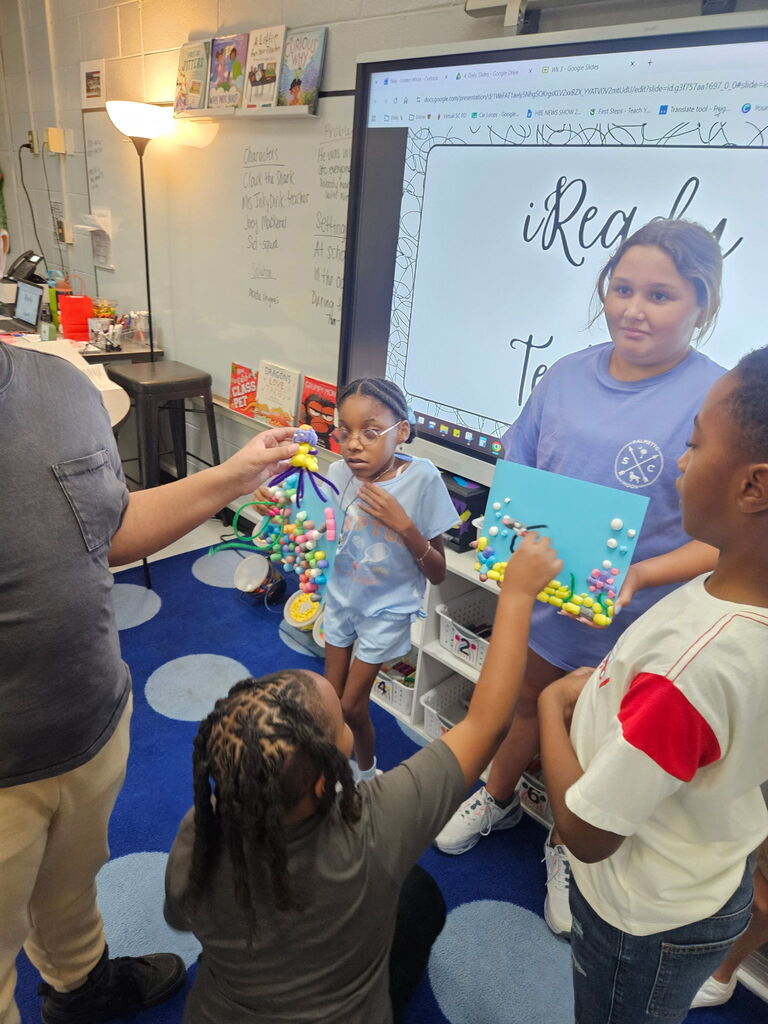 Classroom with a group of children and an adult. Two children hold art projects while another holds a poster.